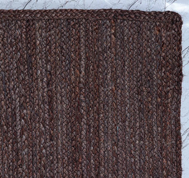 May include: A rectangular area rug featuring a dark brown, braided jute construction. The rug displays a consistent, tight weave pattern with a thick, reinforced border edge that frames the textured surface, showcasing natural fiber variations.