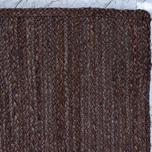 Op de afbeelding: Een rechthoekig kleed met een donkerbruine, gevlochten jute constructie en een dikke, geweven rand. Het oppervlak vertoont een consistent, gestructureerd patroon van verticale, strak verweven vezels die een uniform en aards uiterlijk cre&euml;ren.