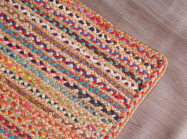 May include: A corner of a handwoven jute rug with a braided design. The rug features a natural tan base with colorful woven stripes in red, blue, green, and yellow. The rug is placed on a light brown wooden floor.