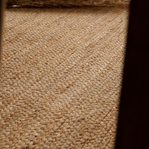 May include: Close-up of a woven jute rug with a natural, light brown color. The rug has a textured surface with visible fibers and a diagonal pattern. The jute material gives a rustic and organic feel.