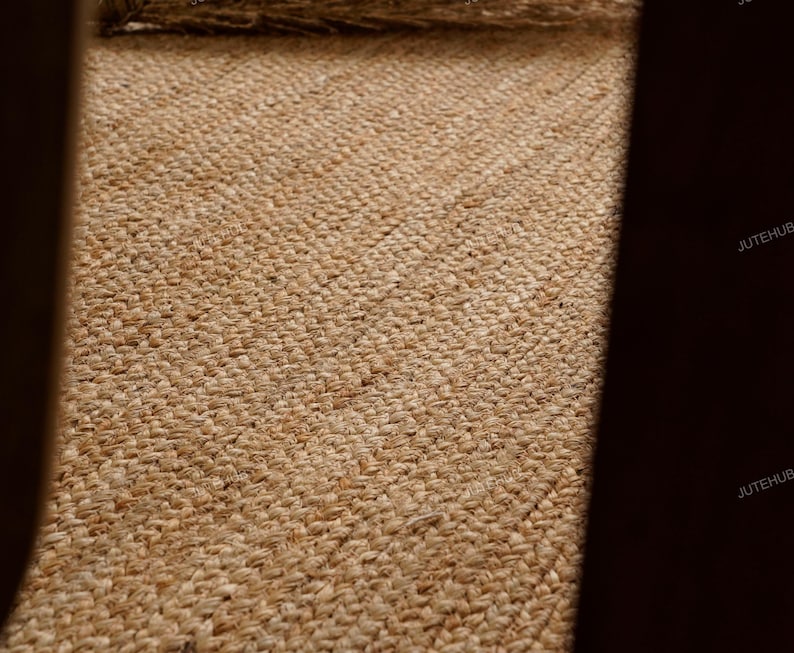 May include: Close-up of a woven jute rug with a natural, light brown color. The rug has a textured surface with visible fibers and a diagonal pattern. The jute material gives a rustic and organic feel.