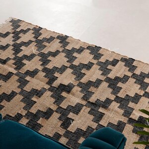 May include: A rectangular rug featuring a geometric design in black and tan. The rug is made from a woven material and has a fringed edge. It's placed on a light-coloured floor, with a teal sofa visible in the foreground.