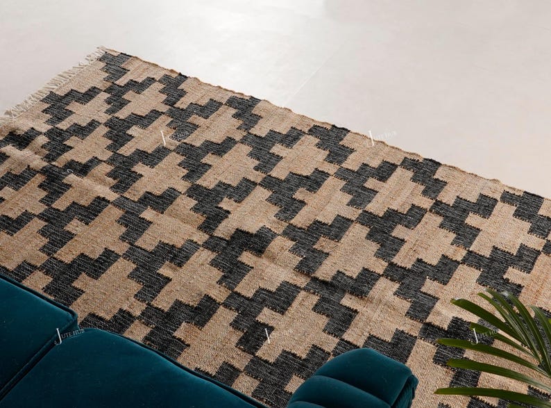 May include: A rectangular rug featuring a geometric design in black and tan. The rug is made from a woven material and has a fringed edge. It's placed on a light-coloured floor, with a teal sofa visible in the foreground.
