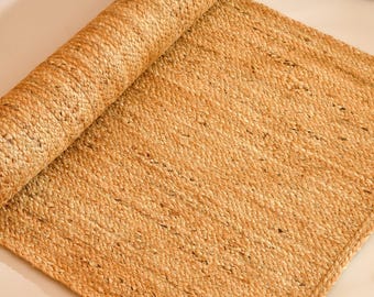 Puede incluir: Una alfombra rectangular hecha de fibras de yute natural de color tostado, con una textura trenzada de tejido apretado. La alfombra está parcialmente enrollada en un extremo, mostrando un patrón de tejido denso y uniforme en tonos tierra.