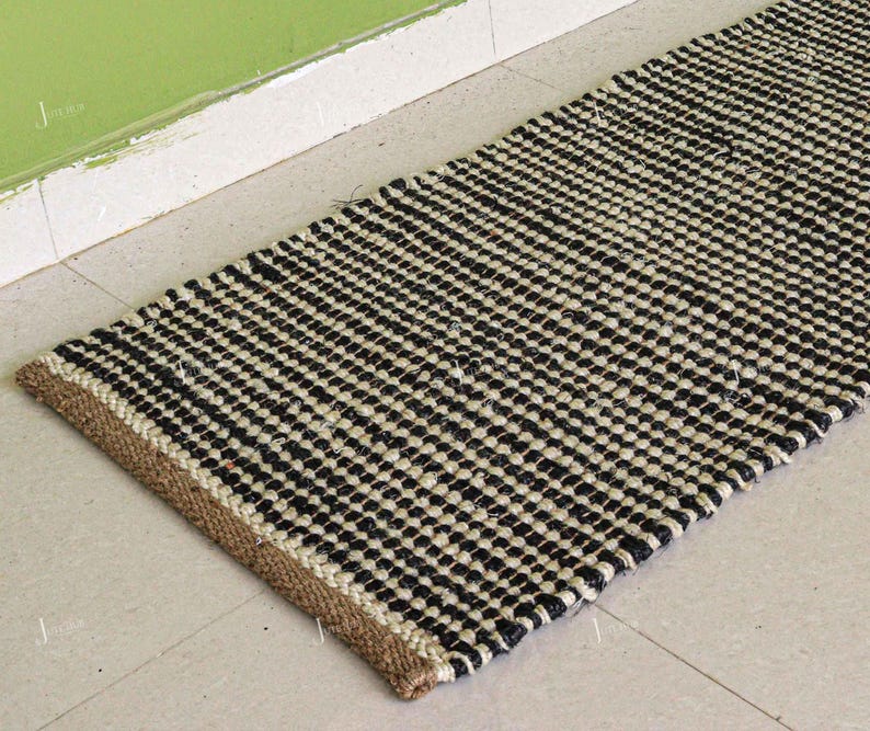 May include: A rectangular black and white woven rug with a repeating pattern of small squares. The rug has a natural fiber border in a tan color. The rug is placed on a light-colored tile floor.
