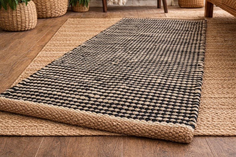 May include: A rectangular area rug featuring a woven texture with a repeating pattern of black and natural tan fibers. The rug has a thick, braided border in a solid natural tan color, creating a distinct contrast against the central pattern.