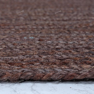May include: A dark brown area rug featuring a thick, braided jute fiber construction with a visible woven texture. The rug has a rounded edge and a repeating watermark text that reads JUTEHUB across the surface of the dark brown material.