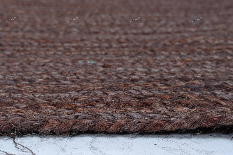 May include: A dark brown area rug featuring a thick, braided jute fiber construction with a visible woven texture. The rug has a rounded edge and a repeating watermark text that reads JUTEHUB across the surface of the dark brown material.