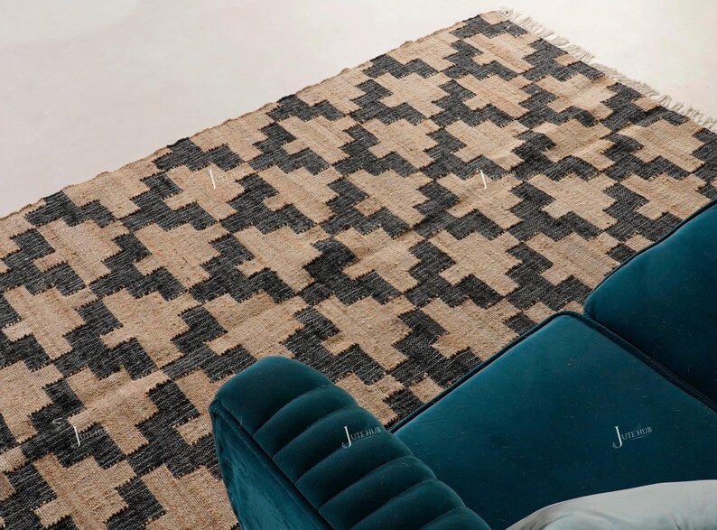 May include: A rectangular jute rug with a geometric pattern in black and tan. The rug has a fringed edge and is next to a teal sofa. The rug's design is a repeating series of angular shapes.