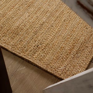 May include: A rectangular jute rug with a woven texture. The rug is a natural tan color and appears to be handmade. The rug is placed on a wooden floor, with a portion of a chair visible in the frame. The rug is a home decor item.