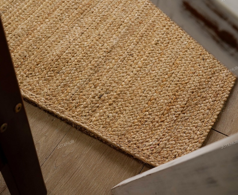 May include: A rectangular jute rug with a woven texture. The rug is a natural tan color and appears to be handmade. The rug is placed on a wooden floor, with a portion of a chair visible in the frame. The rug is a home decor item.