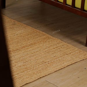 May include: A rectangular, natural-toned jute rug with a woven texture. The rug is placed on a light-colored wooden floor, with a dark wooden bed frame visible in the background. The rug's color is a warm, earthy tone.