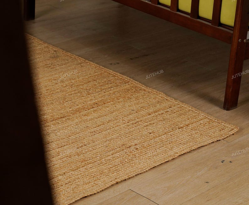 May include: A rectangular, natural-toned jute rug with a woven texture. The rug is placed on a light-colored wooden floor, with a dark wooden bed frame visible in the background. The rug's color is a warm, earthy tone.