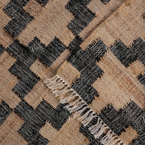 May include: A woven rug with a geometric pattern. The rug features a repeating design of black and tan shapes. The edges of the rug have a fringe detail. The rug is made of a natural fibre.