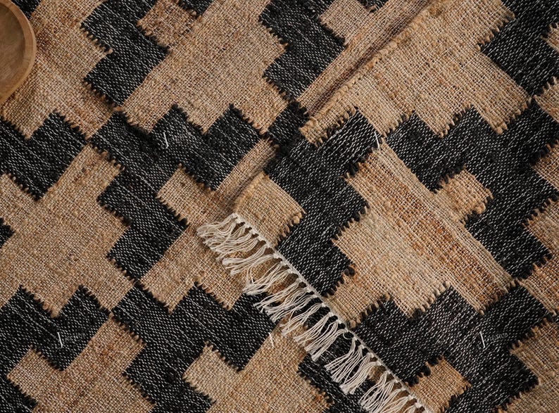 May include: A woven rug with a geometric pattern. The rug features a repeating design of black and tan shapes. The edges of the rug have a fringe detail. The rug is made of a natural fibre.