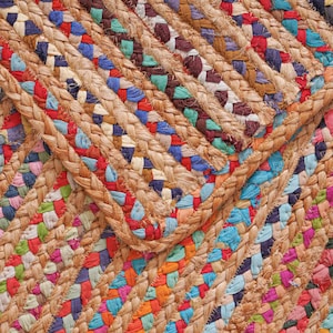 May include: Close-up of a colorful braided jute rug. The rug features a mix of vibrant colors including red, blue, green, and pink, woven into a textured pattern. The natural jute fibers provide a warm, earthy tone.