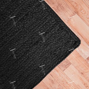 May include: A rectangular area rug featuring a solid black colour and a braided texture. The surface of the rug is covered with a repeating white watermark logo that reads JUTE HUB in a vertical orientation across the entire black fabric material.
