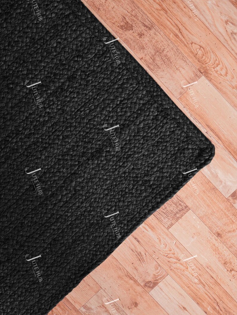May include: A rectangular area rug featuring a solid black colour and a braided texture. The surface of the rug is covered with a repeating white watermark logo that reads JUTE HUB in a vertical orientation across the entire black fabric material.