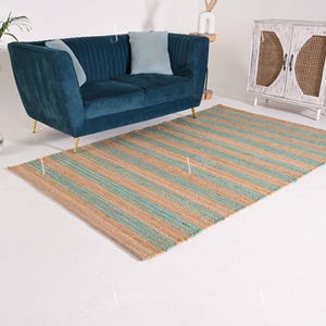Op de afbeelding: Een rechthoekig jute vloerkleed met afwisselende strepen in beige en turquoise. Het vloerkleed ligt op een witte vloer, met een teal fluwelen bank en een witte kast op de achtergrond. Het vloerkleed is een woondecoratie-artikel.