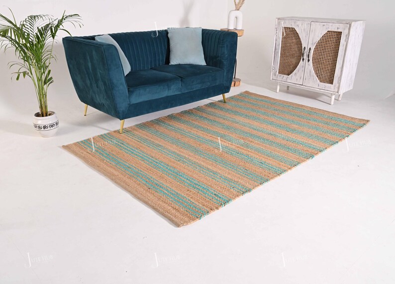 Op de afbeelding: Een rechthoekig jute vloerkleed met afwisselende strepen in beige en turquoise. Het vloerkleed ligt op een witte vloer, met een teal fluwelen bank en een witte kast op de achtergrond. Het vloerkleed is een woondecoratie-artikel.