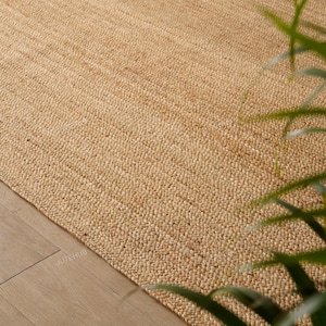 May include: A close-up of a natural jute rug with a woven texture. The rug is a light tan color and appears to be handmade. The rug is placed on a light-colored wooden floor.