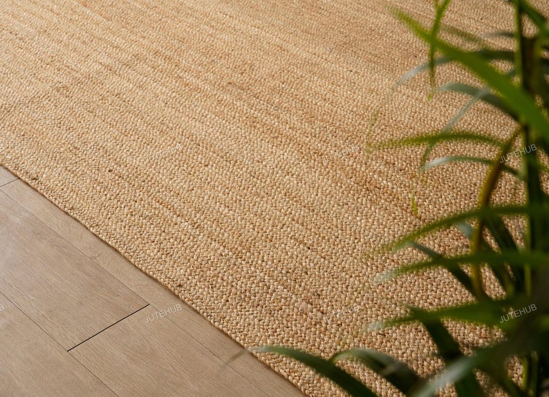 May include: A close-up of a natural jute rug with a woven texture. The rug is a light tan color and appears to be handmade. The rug is placed on a light-colored wooden floor.