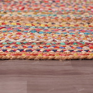 May include: Close-up of a colorful braided jute rug. The rug features a mix of vibrant colors including red, blue, yellow, and green, woven together in a textured pattern. The rug is placed on a wooden floor.