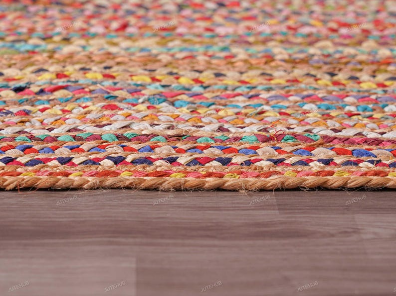 May include: Close-up of a colorful braided jute rug. The rug features a mix of vibrant colors including red, blue, yellow, and green, woven together in a textured pattern. The rug is placed on a wooden floor.