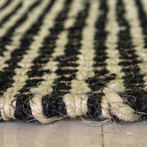 May include: A woven rug featuring a repeating striped pattern of natural beige and black fibres. The texture is coarse and thick, with visible braided strands creating a durable surface. The design consists of alternating vertical stripes.