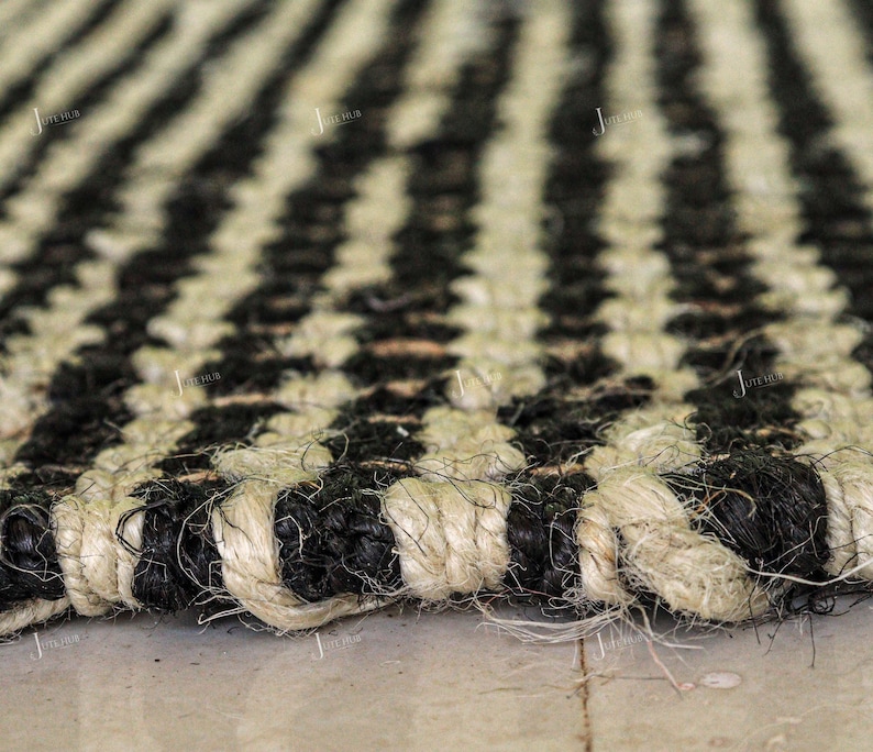 May include: A woven rug featuring a repeating striped pattern of natural beige and black fibres. The texture is coarse and thick, with visible braided strands creating a durable surface. The design consists of alternating vertical stripes.