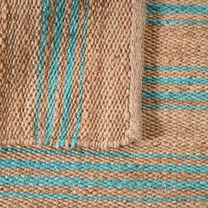 Op de afbeelding: Close-up van een geweven jute vloerkleed met afwisselende strepen in natuurlijk bruin en turquoise blauw. Het vloerkleed heeft een getextureerd oppervlak en een opgerolde rand, die de weefsel en kleurvariaties van het materiaal laat zien.
