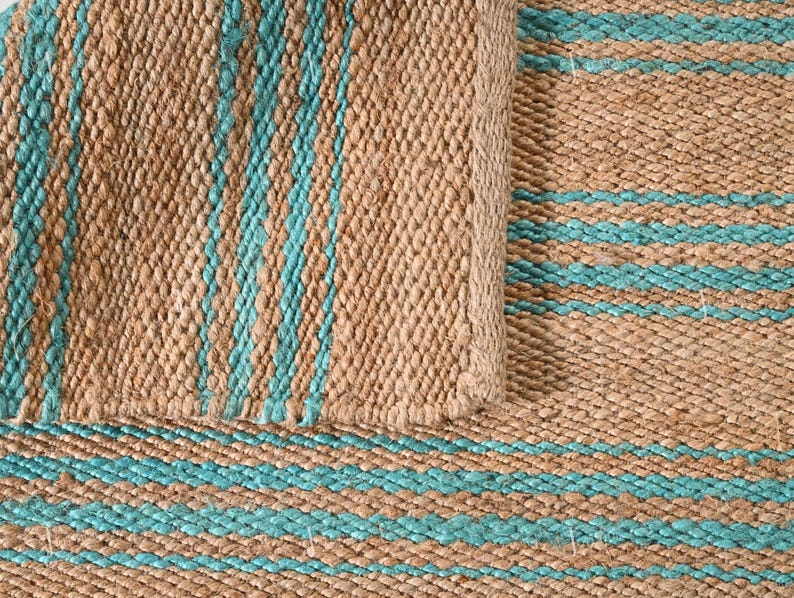 Op de afbeelding: Close-up van een geweven jute vloerkleed met afwisselende strepen in natuurlijk bruin en turquoise blauw. Het vloerkleed heeft een getextureerd oppervlak en een opgerolde rand, die de weefsel en kleurvariaties van het materiaal laat zien.