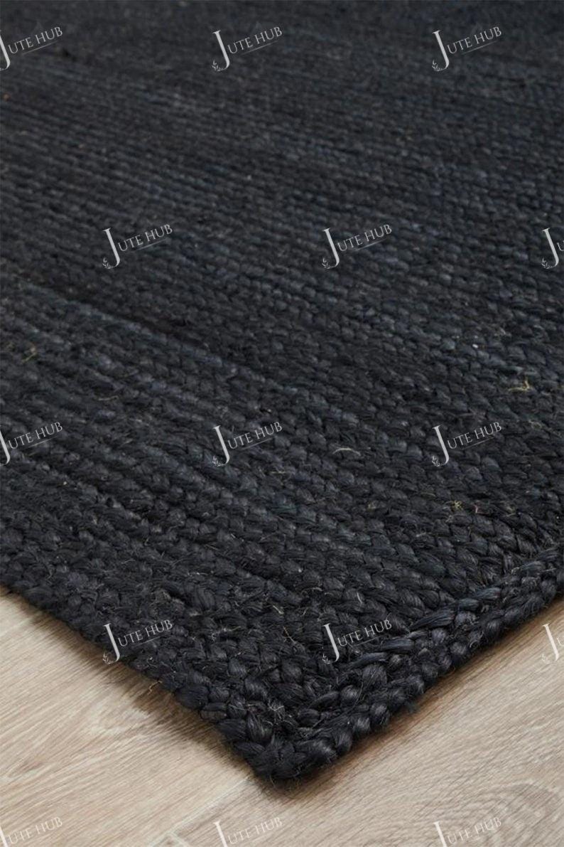 May include: Close-up of a black jute rug with a woven texture. The rug's corner is visible, showcasing the thick, durable material. The dark color and natural fibers suggest a rustic or modern home decor item.