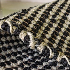 May include: A woven rug featuring a high-contrast checkerboard pattern in black and off-white. The texture is coarse and fibrous, with a thick, braided edge finish. The repeating square design is visible across the surface and the folded corner.