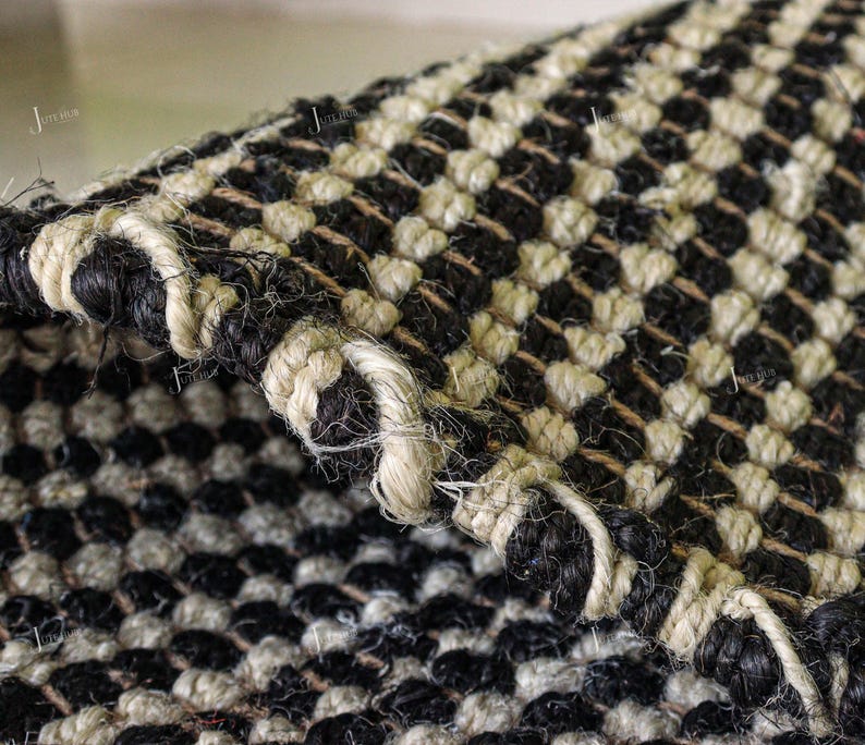May include: A woven rug featuring a high-contrast checkerboard pattern in black and off-white. The texture is coarse and fibrous, with a thick, braided edge finish. The repeating square design is visible across the surface and the folded corner.