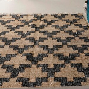 May include: Rectangular rug featuring a geometric pattern in black and beige. The rug displays a repeating design of interlocking shapes. It is made from a natural fibre and has a fringed edge. The rug is placed on a light-coloured floor.