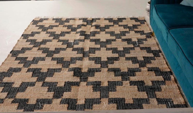 May include: Rectangular rug featuring a geometric pattern in black and beige. The rug displays a repeating design of interlocking shapes. It is made from a natural fibre and has a fringed edge. The rug is placed on a light-coloured floor.