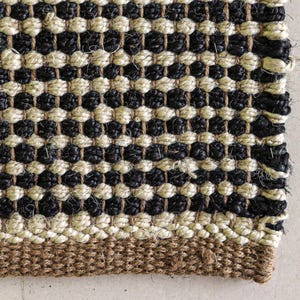 May include: A woven rug featuring a repeating pattern of alternating horizontal rows of black and cream-coloured thick fibres. The bottom edge displays a solid, wider band of natural brown woven material, creating a textured, durable surface.