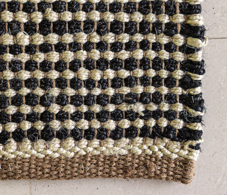 May include: A woven rug featuring a repeating pattern of alternating horizontal rows of black and cream-coloured thick fibres. The bottom edge displays a solid, wider band of natural brown woven material, creating a textured, durable surface.