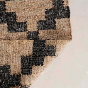 May include: Close-up of a jute rug with a geometric design. The rug has a natural tan base with black square and rectangular shapes. The edge is folded over, showing the underside of the rug.