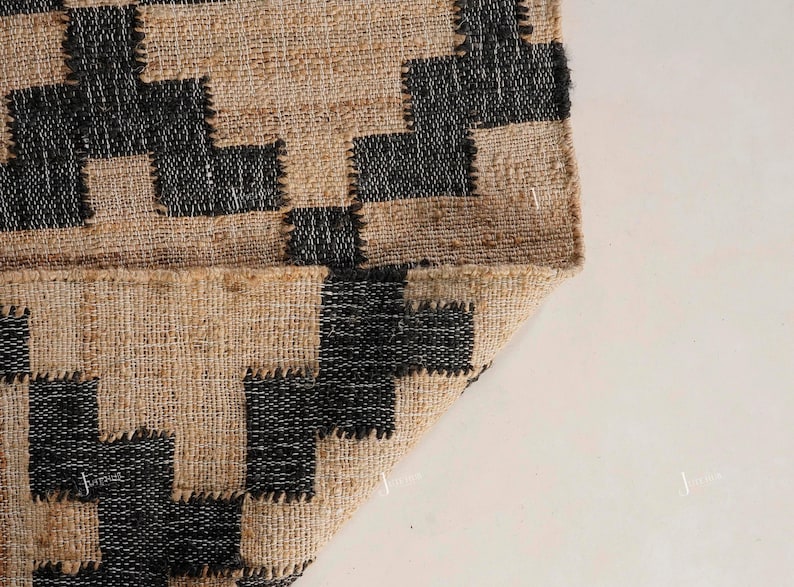 May include: Close-up of a jute rug with a geometric design. The rug has a natural tan base with black square and rectangular shapes. The edge is folded over, showing the underside of the rug.