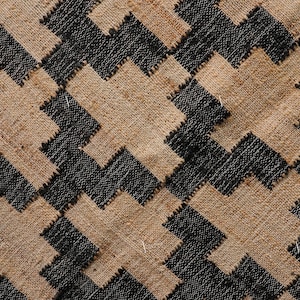 May include: Close-up of a woven textile with a repeating geometric pattern. The design features interlocking shapes in black and tan, creating a textured surface. The weave appears to be made of natural fibres.