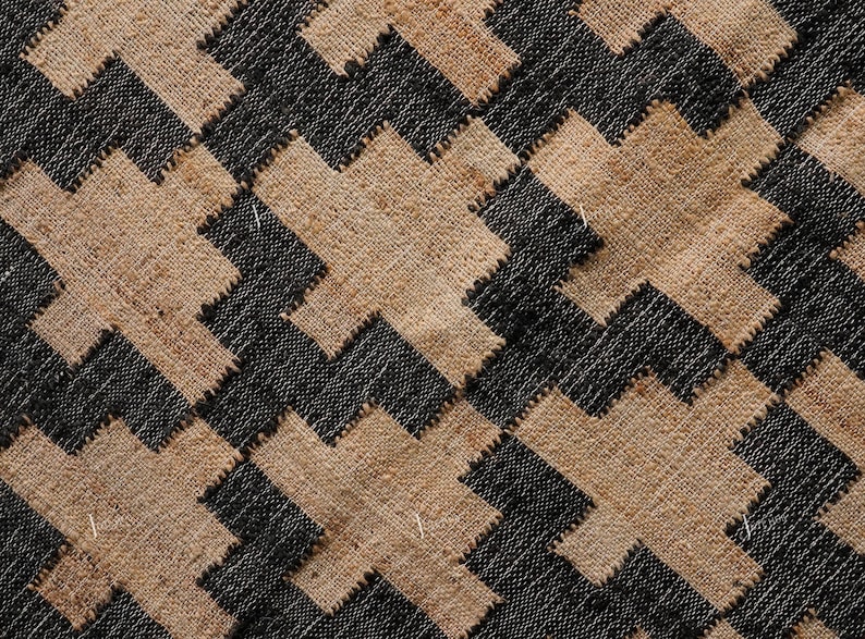 May include: Close-up of a woven textile with a repeating geometric pattern. The design features interlocking shapes in black and tan, creating a textured surface. The weave appears to be made of natural fibres.