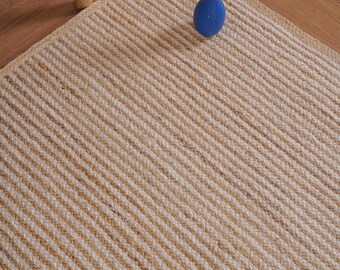 Puede incluir: Una alfombra rectangular con textura trenzada y rayas alternas de yute color canela natural y fibras de color blanco roto. La alfombra presenta una construcción tejida gruesa con un patrón consistente en toda su superficie.
