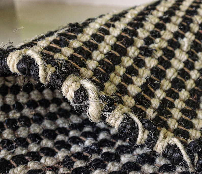 May include: A woven rug featuring a repeating checkerboard pattern of black and off-white natural fibers. The thick, textured material is folded over to reveal the dense, knotted construction and the sturdy, braided edge of the textile.