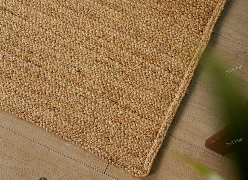 May include: Close-up of a natural jute rug with a woven texture. The rug has a light brown color and a braided edge. The corner of the rug is visible, showing the texture and construction. The rug is on a wooden floor.