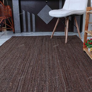 May include: A rectangular area rug featuring a dark brown, braided jute texture with a consistent woven pattern. The rug lies flat on a light-colored floor, positioned in front of a white chair and a wooden shelving unit containing small potted plants.