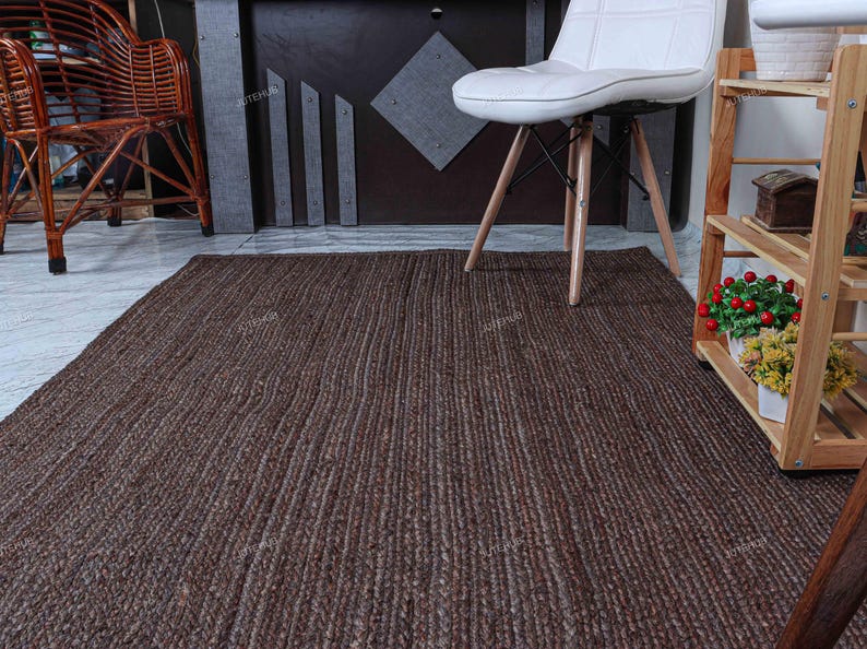 May include: A rectangular area rug featuring a dark brown, braided jute texture with a consistent woven pattern. The rug lies flat on a light-colored floor, positioned in front of a white chair and a wooden shelving unit containing small potted plants.