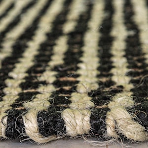 May include: A woven rug featuring a repeating striped pattern of thick, natural tan jute fibers and dyed black fibers. The texture is coarse and braided, with the word JUTEHUB printed in small, repeating text across the surface of the rug.