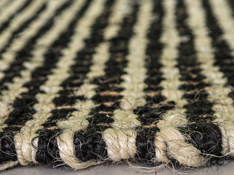 May include: A woven rug featuring a repeating striped pattern of thick, natural tan jute fibers and dyed black fibers. The texture is coarse and braided, with the word JUTEHUB printed in small, repeating text across the surface of the rug.